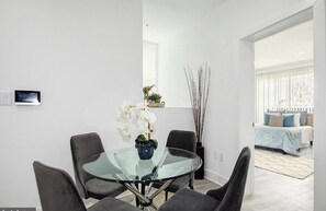 Un comedor moderno con una mesa de vidrio y sillas grises tapizadas, una pequeña planta en maceta y un jarrón decorativo con flores blancas.