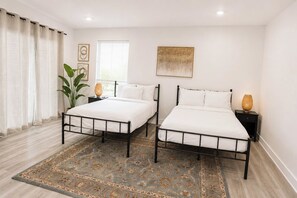 Un dormitorio con dos camas, un ventanal con persianas, un tapete decorativo y una planta.