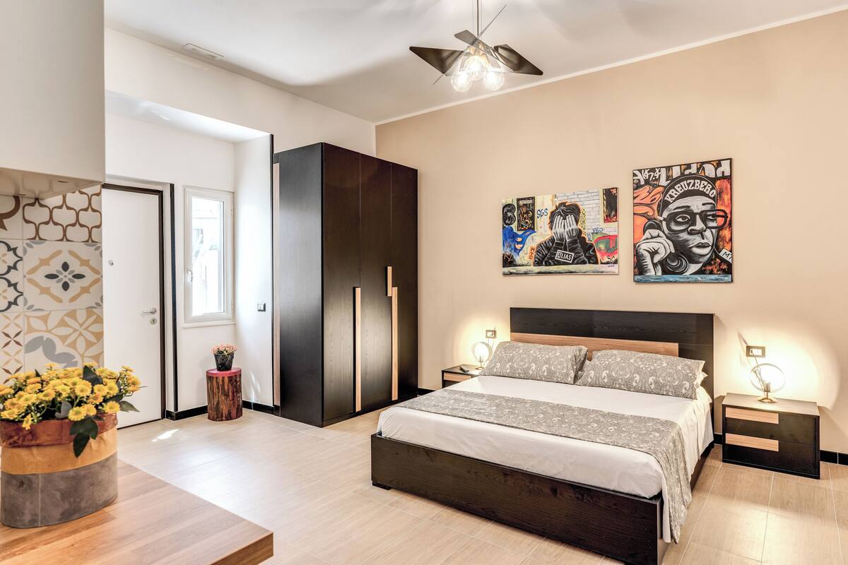 A modern bedroom with a large bed, a wardrobe, and a ceiling fan. There are two wall art pieces and a small table with flowers.