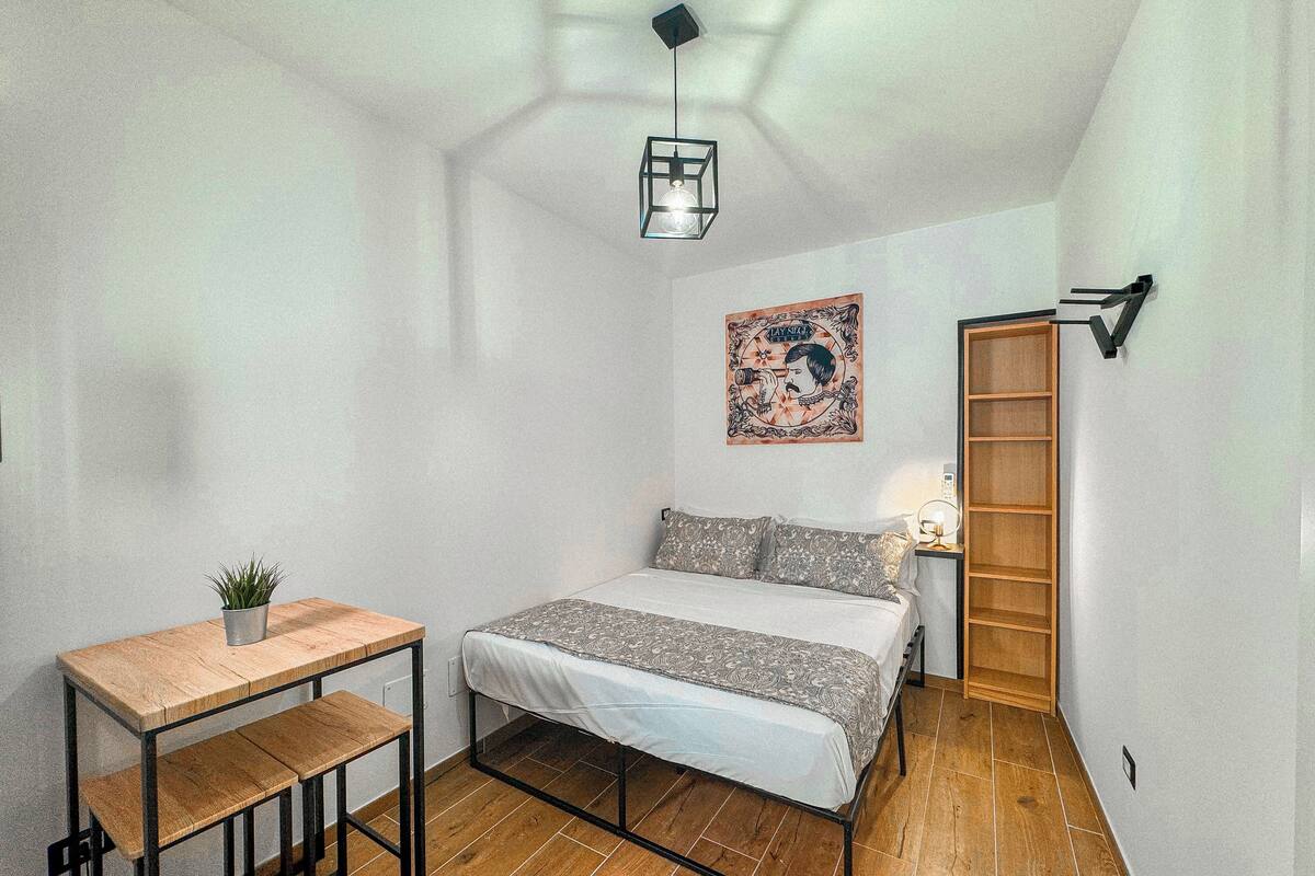 A bedroom with a bed, a small table, a potted plant, a wall-mounted shelf, and a door.