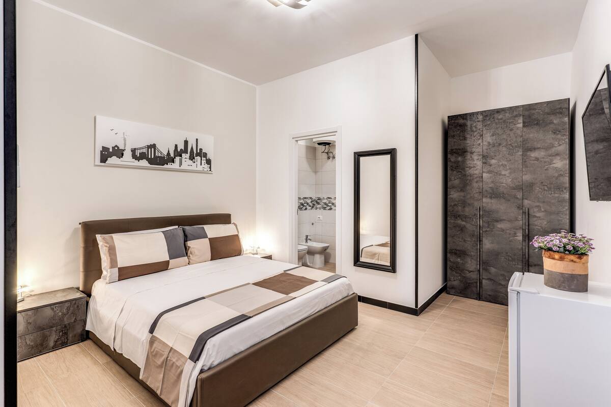 A modern bedroom with a large bed, a cityscape artwork, a mirror, and a wardrobe.