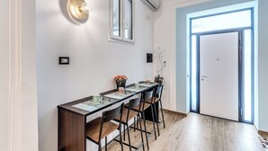 A dining area with a long table, bar stools, wall-mounted light fixtures, and a door leading to a bright room. - The Quarters Naples Apartment (Naples)