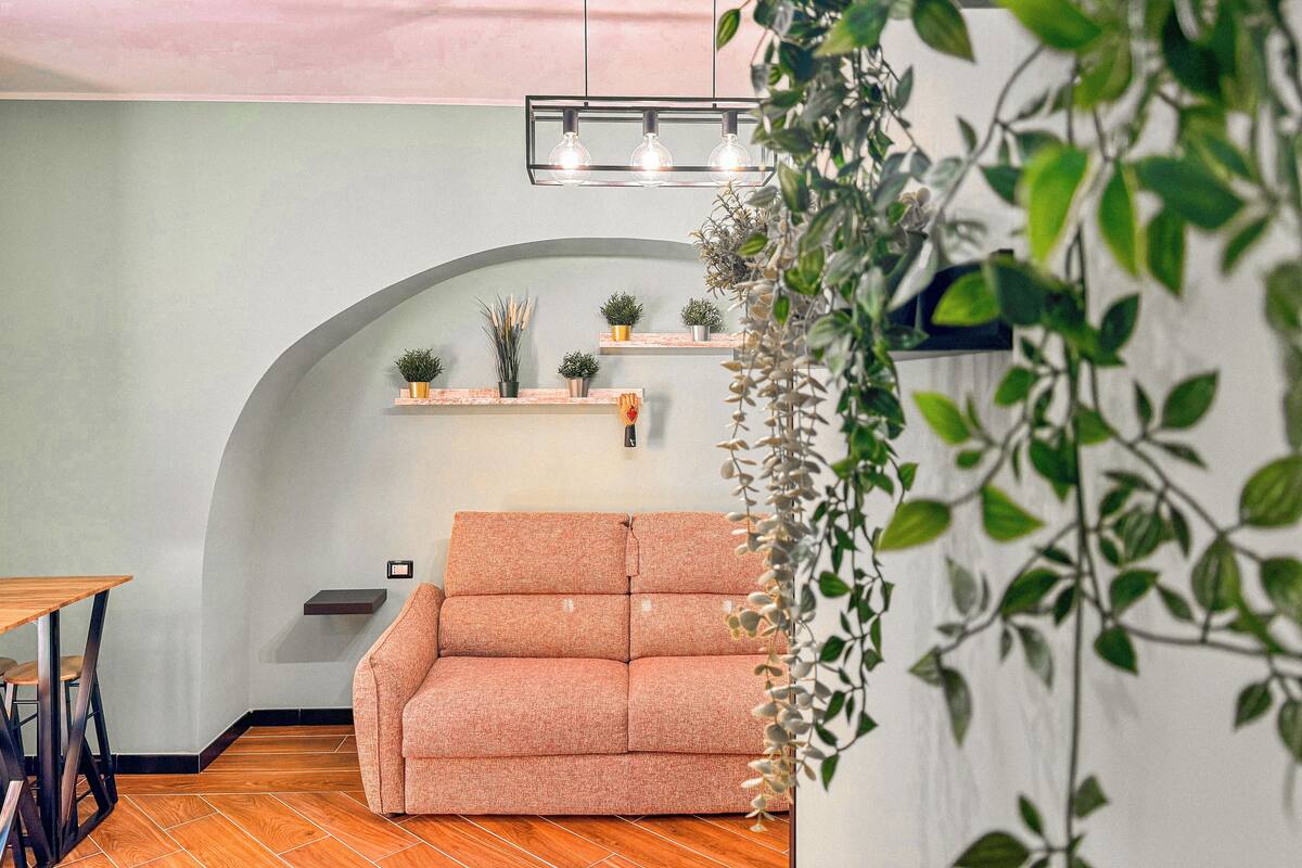 A cozy living area with a sofa, wooden flooring, a ceiling light fixture, and wall-mounted shelves with plants.