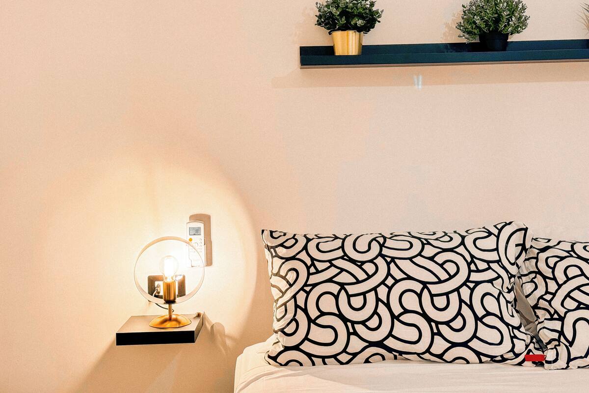 A bedroom with a bed, a bedside lamp, a wall clock, and two potted plants on a shelf above the bed.