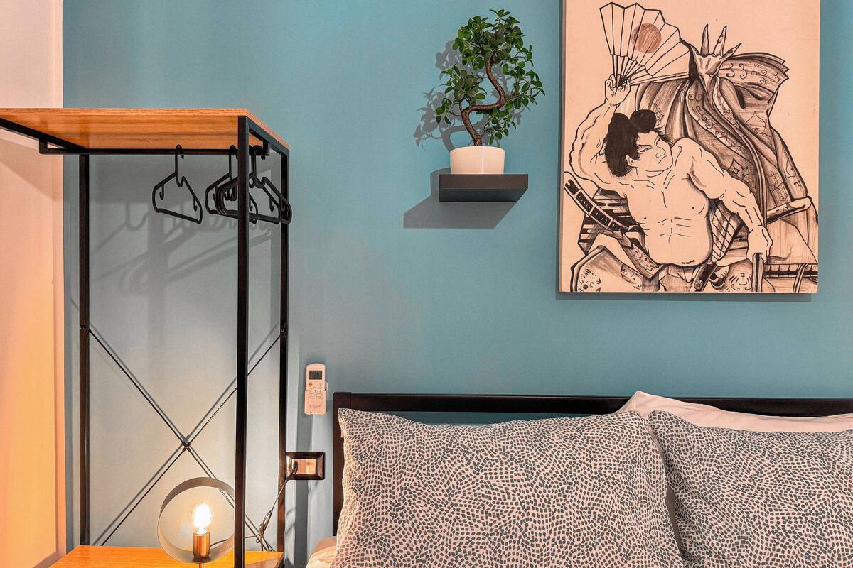A bedroom with a bed, a bedside table with a lamp, a hanging plant, and a wall-mounted shelf with a plant and a framed artwork.