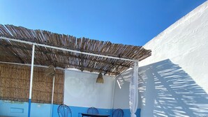 Un espacio al aire libre cubierto con piscina, suelo de baldosas azules y blancas, una mesa con sillas y techo de paja.