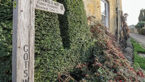 Et træskilt med teksten 'COTSWOLD WAY' og 'Broadway Tower 1½' mod en baggrund af en bygning og frodig vegetation.