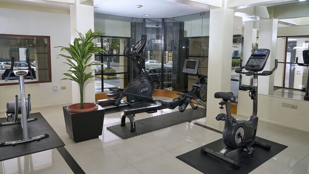 Un gimnasio con diversos equipos de ejercicio, incluyendo una bicicleta estática, una máquina elíptica y una cinta de correr. Hay una planta en maceta y un espejo en la pared.