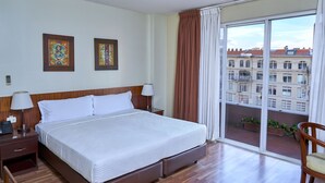 Habitación de hotel con una cama grande, cabecera de madera, dos mesitas de noche con lámparas, vista a edificios a través de un ventanal amplio y un balcón con plantas en macetas.