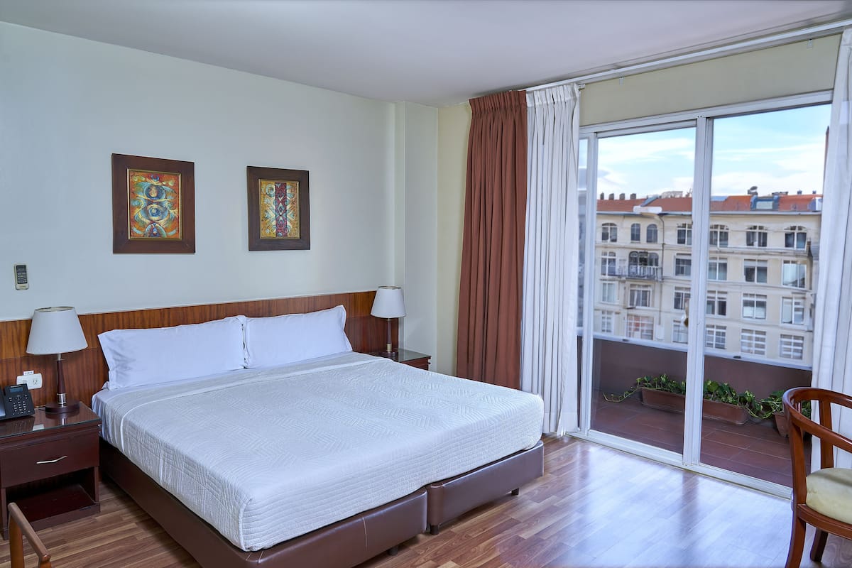 A hotel room with a large bed, wooden headboard, two bedside tables with lamps, a view of buildings through a large window, and a balcony with potted plants.