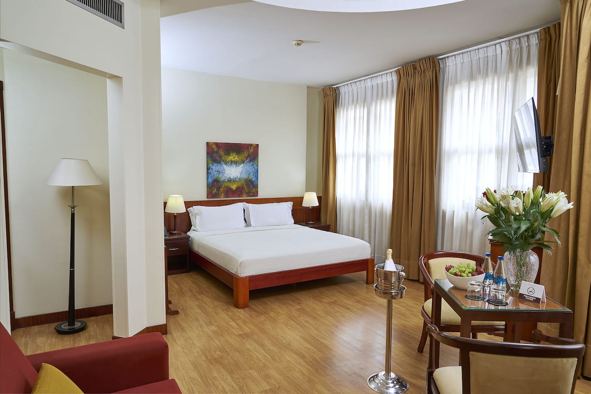 A hotel room with a large bed, a desk with a vase of flowers, a dining table, and a painting on the wall.