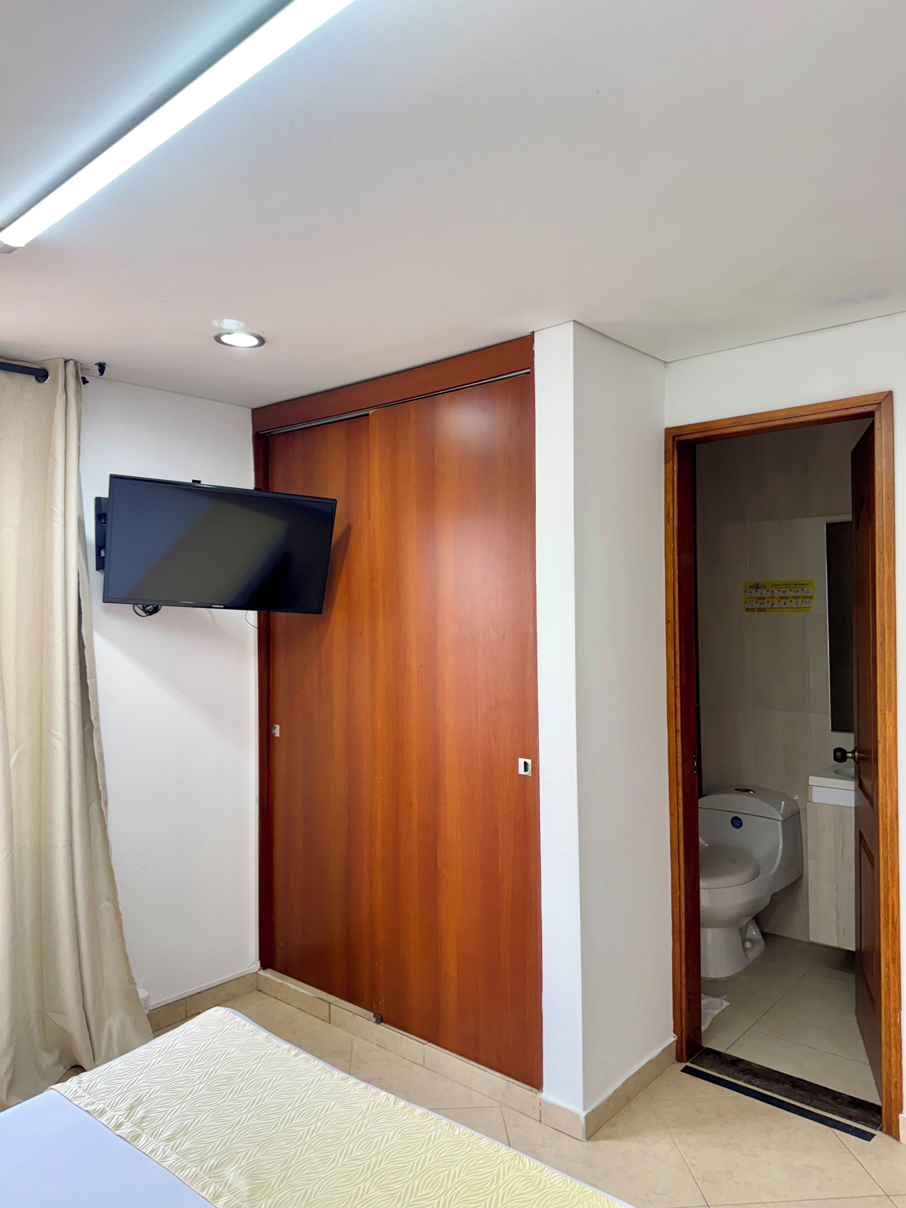 Un dormitorio con un armario de madera, un televisor de pantalla plana montado en la pared, una cama con colcha estampada y un baño visible a través de una puerta abierta.