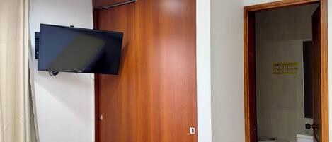 Un dormitorio con un armario de madera, un televisor de pantalla plana montado en la pared, una cama con colcha estampada y un baño visible a través de una puerta abierta.
