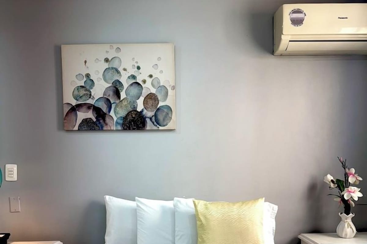 A hotel room with a bed, white bedding, a wall-mounted air conditioner, a bedside table with a vase of flowers, and a painting above the bed.