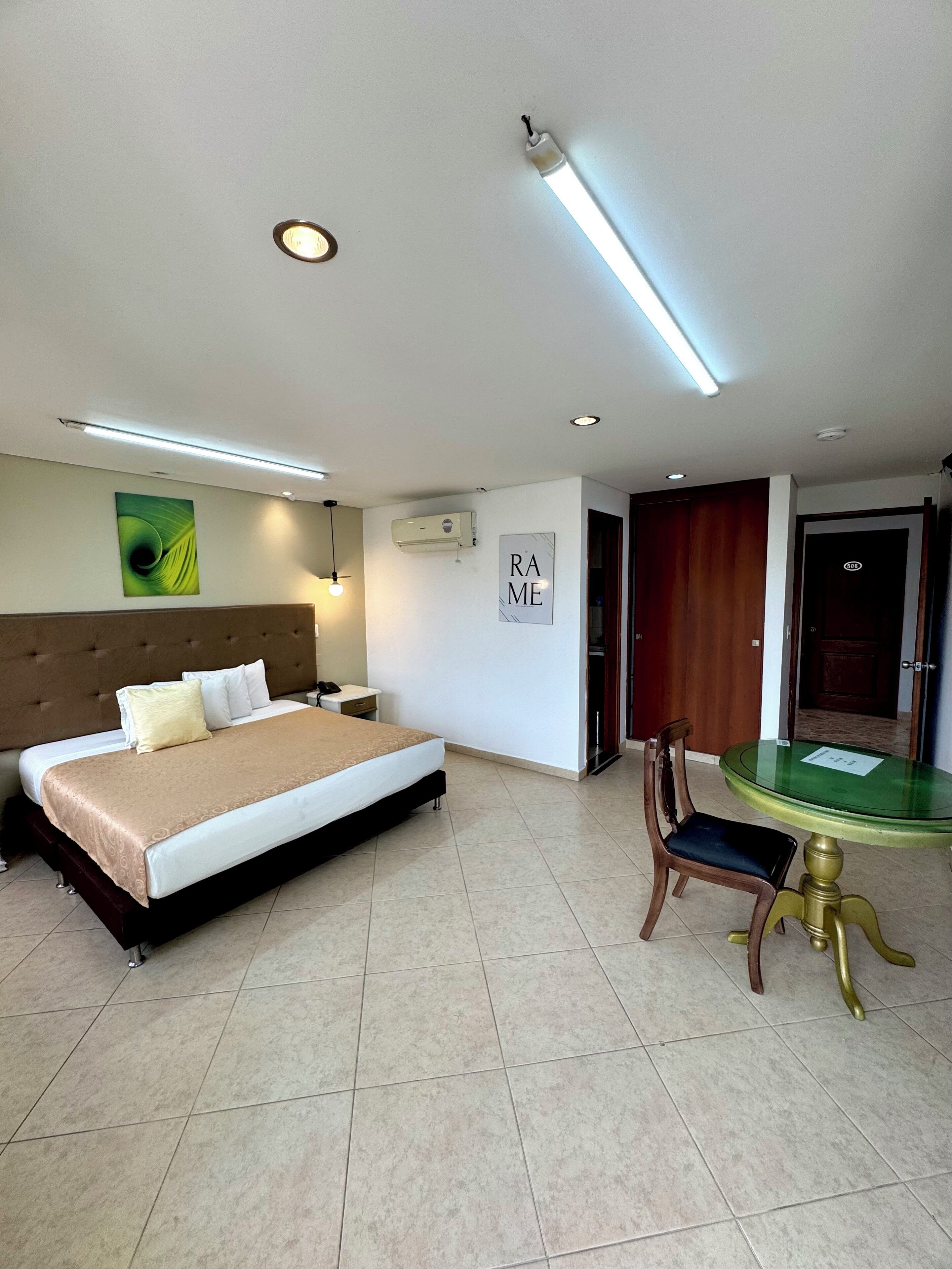 A hotel room with a large bed, a small table with a chair, an air conditioning unit, and a door.