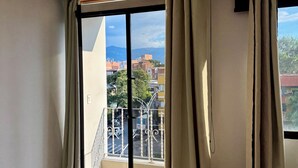 Una habitación con un amplio ventanal corredera que da a un balcón, una lámpara en el techo y vistas a edificios y árboles.