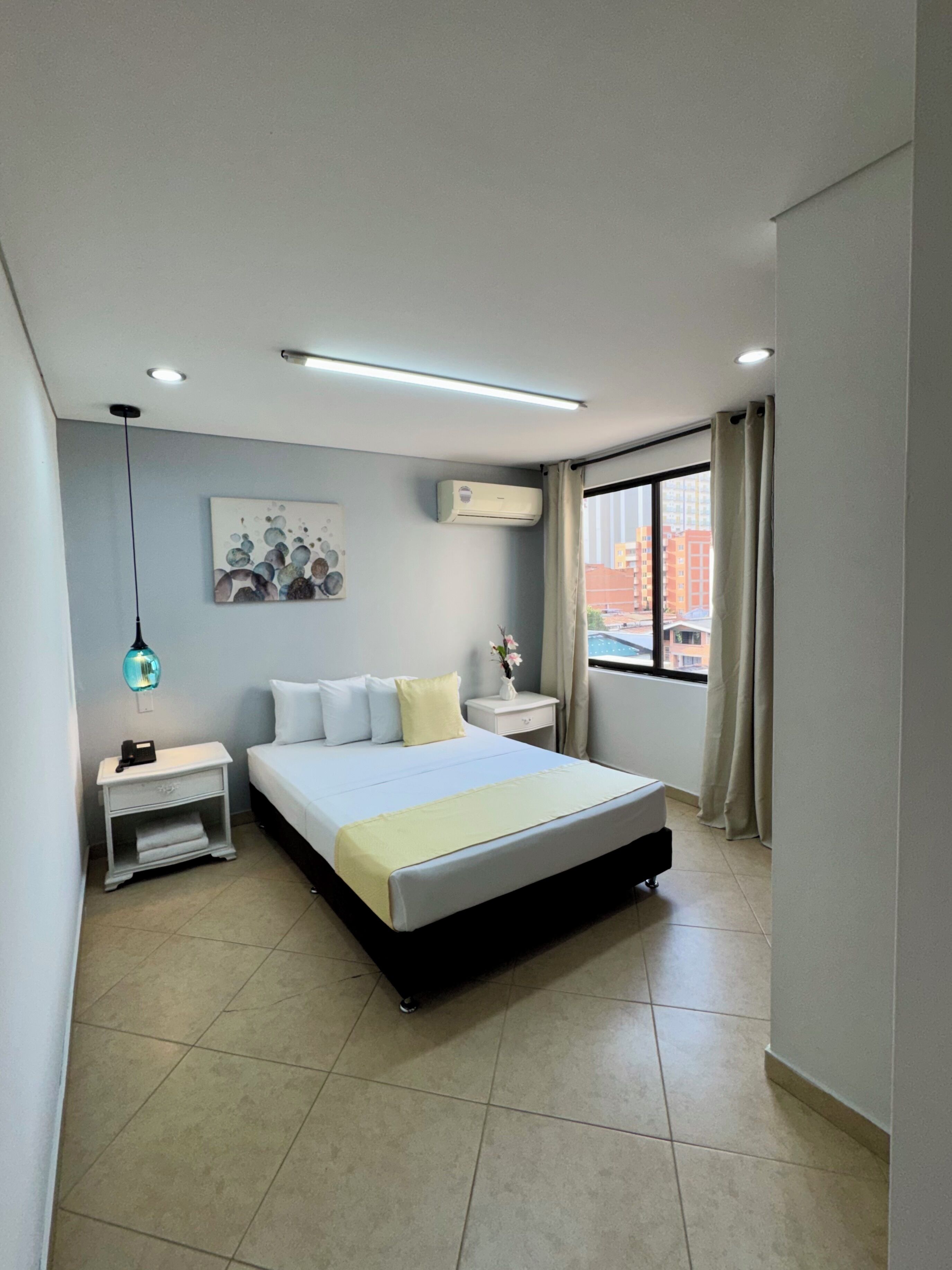 Un dormitorio moderno con cama, mesitas de noche, aire acondicionado, una ventana con cortinas y vistas a la ciudad.