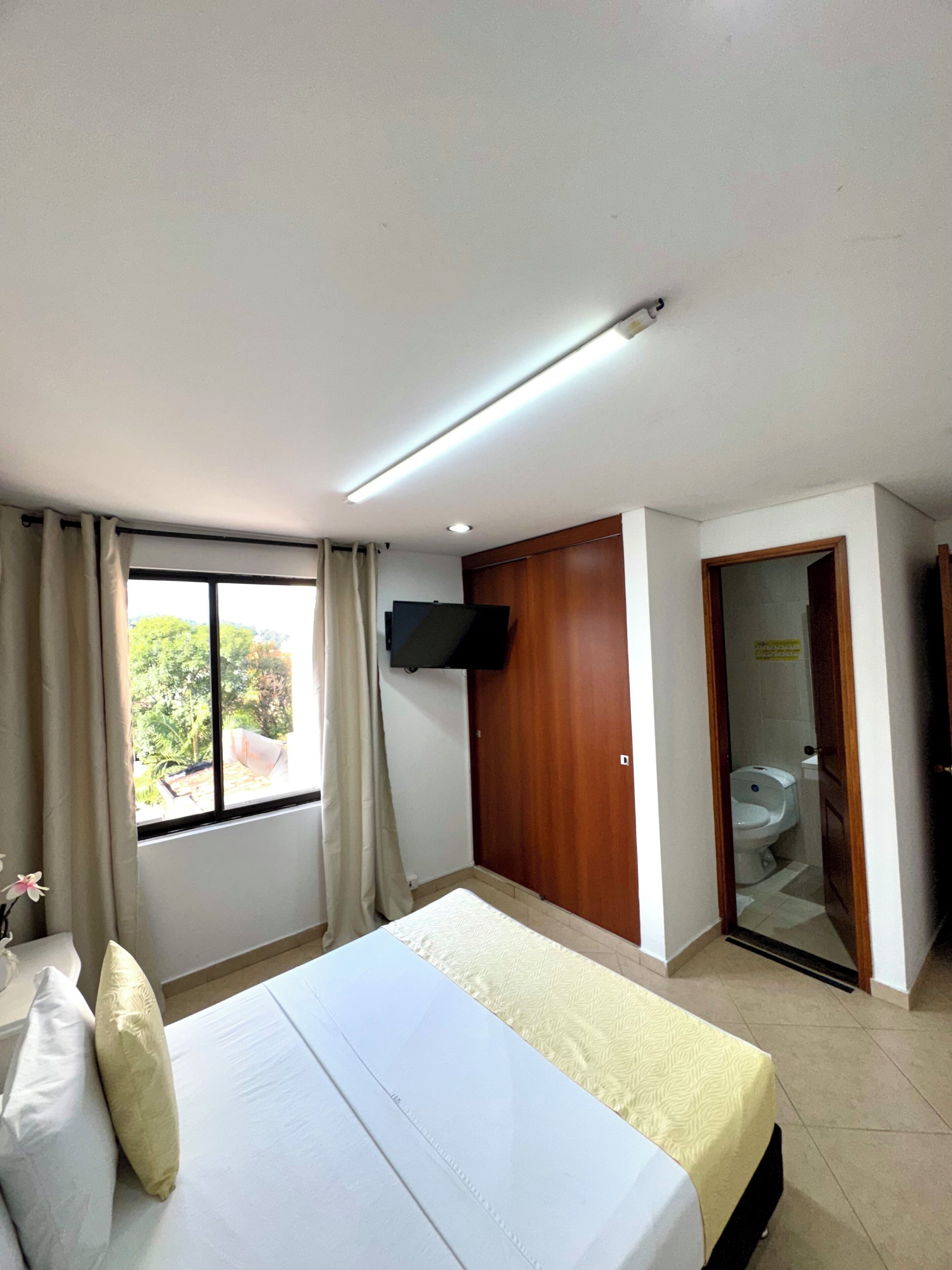 A hotel room with a bed, a TV mounted on the wall, a window with curtains, and a door leading to a bathroom.