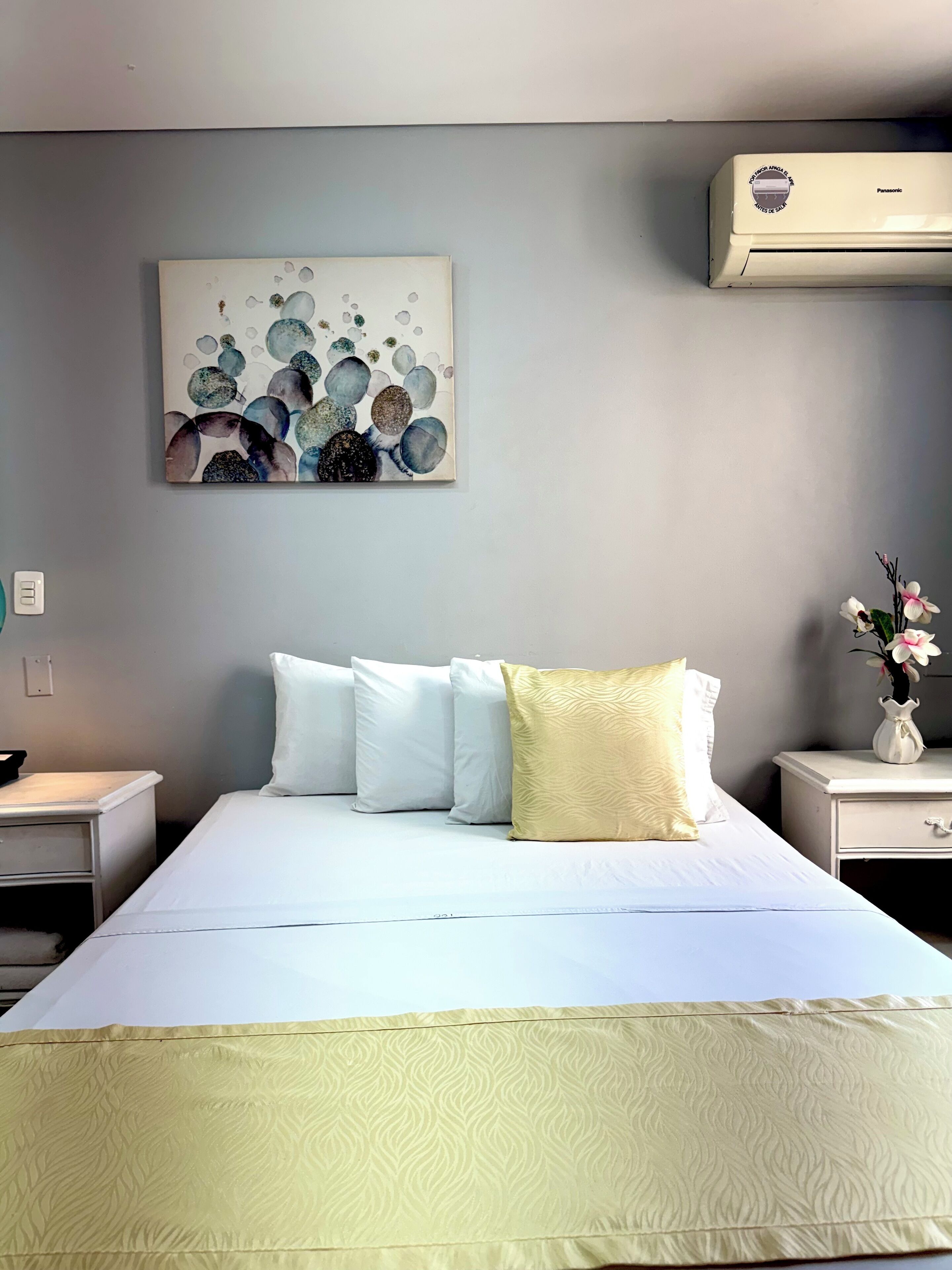Une chambre d’hôtel avec un lit, des oreillers blancs, un oreiller décoratif jaune, un climatiseur fixé au mur et un tableau au-dessus du lit.