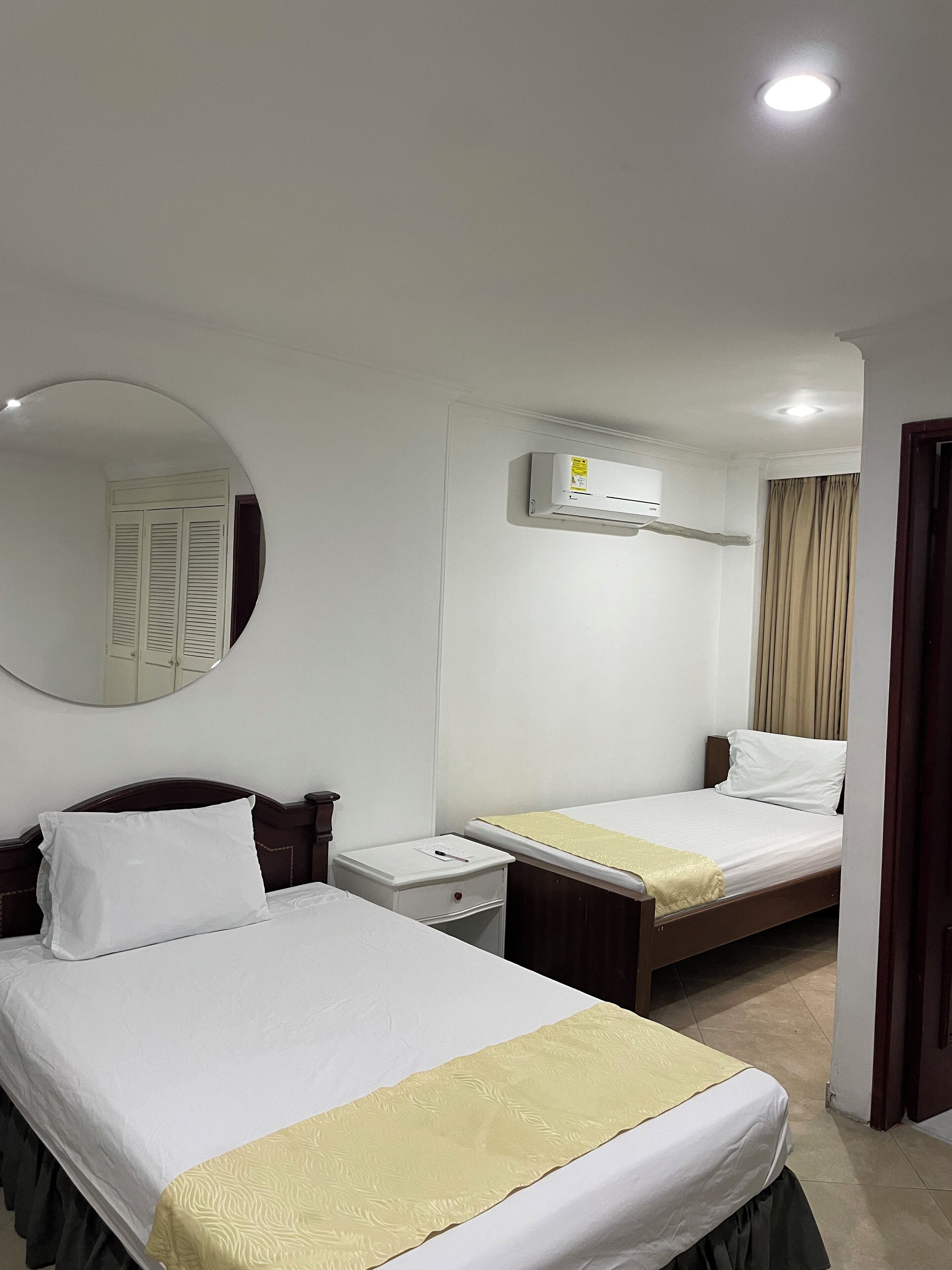 A hotel room with two beds, a round mirror, a ceiling light, and an air conditioning unit.