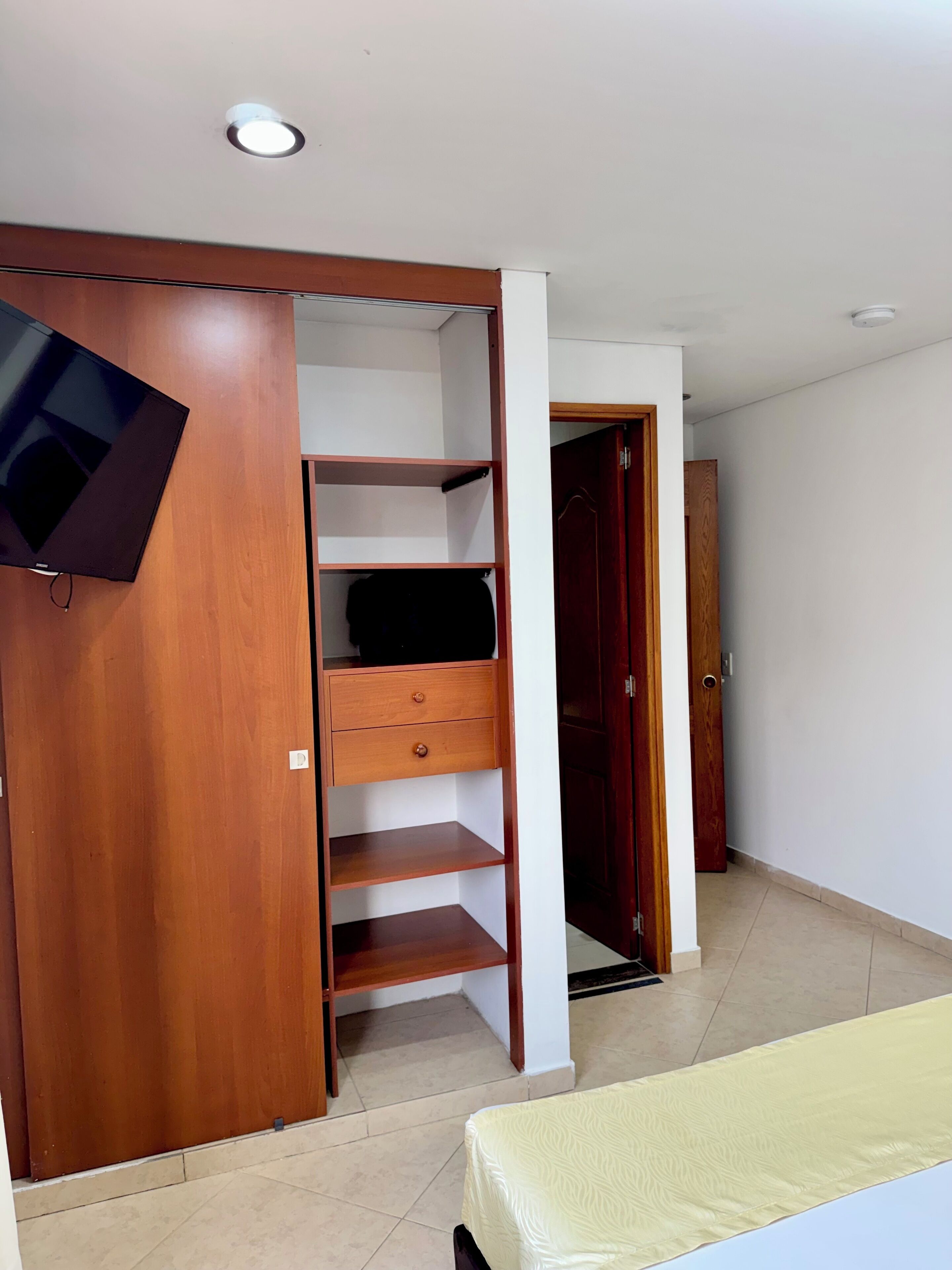 A bedroom with a wooden wardrobe, a bed with a patterned bedspread, and a television mounted on the wall.