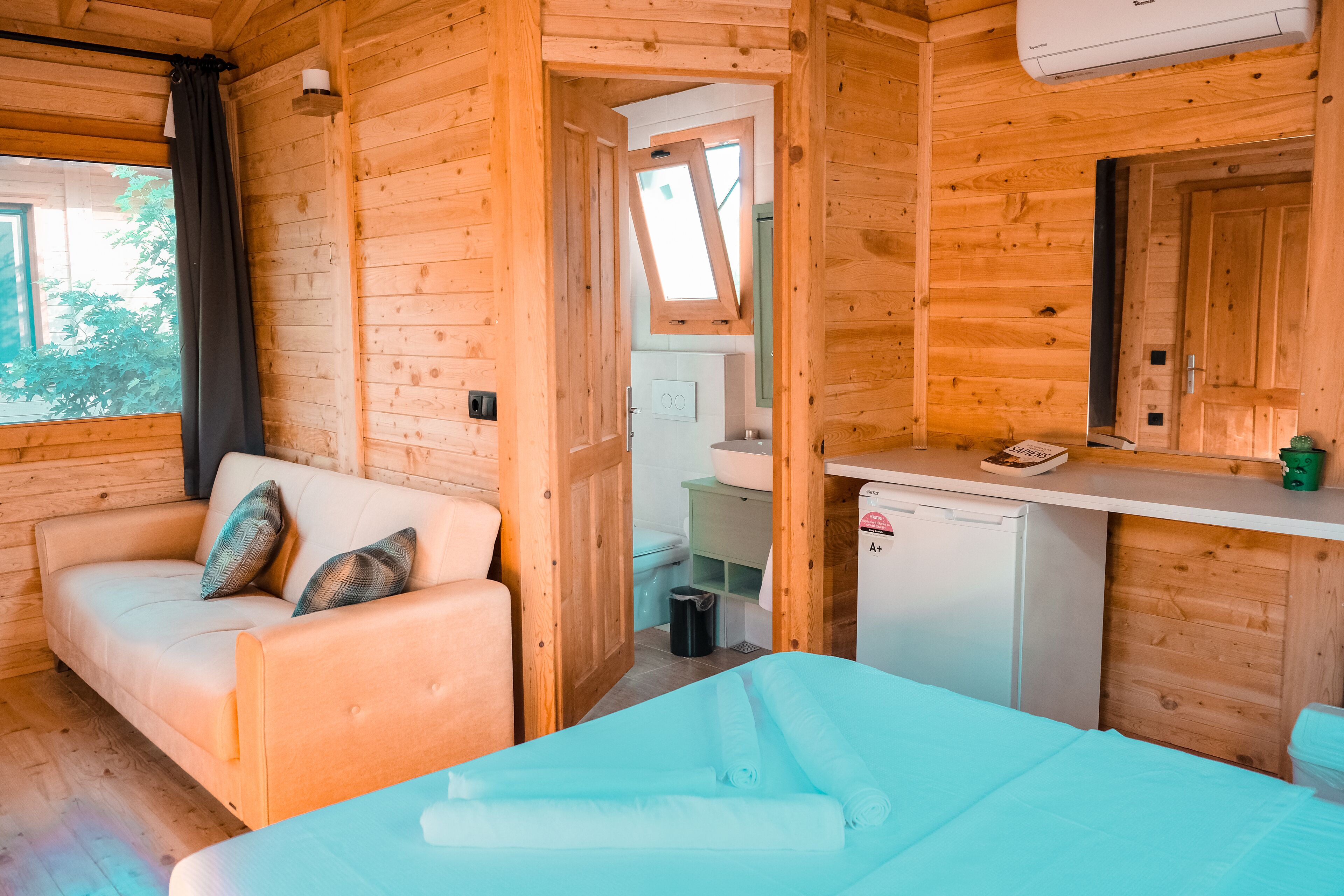 A wooden cabin room with a sofa, a small table, a bed, a kitchenette, and a window.