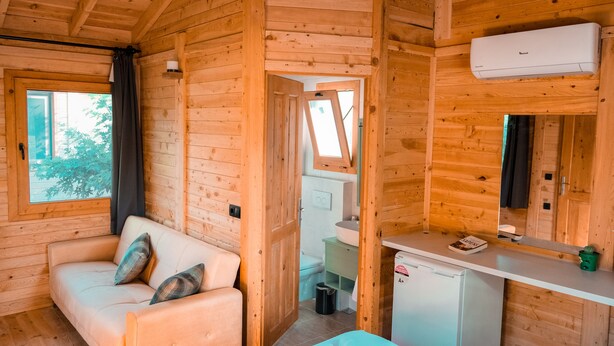 L’intérieur d’un chalet en bois, avec un canapé, une petite cuisine et une salle de bain visible à travers une porte ouverte.