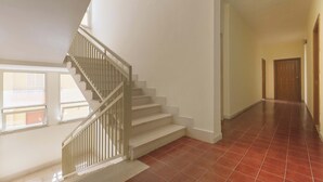 Una escalera con barandillas blancas que conduce a un piso superior, un pasillo con baldosas rojas y una puerta de madera al final.