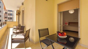 Un balcón con sillas de mimbre, una mesa de vidrio con fruteros y vistas a un dormitorio con cama y mesita de noche.