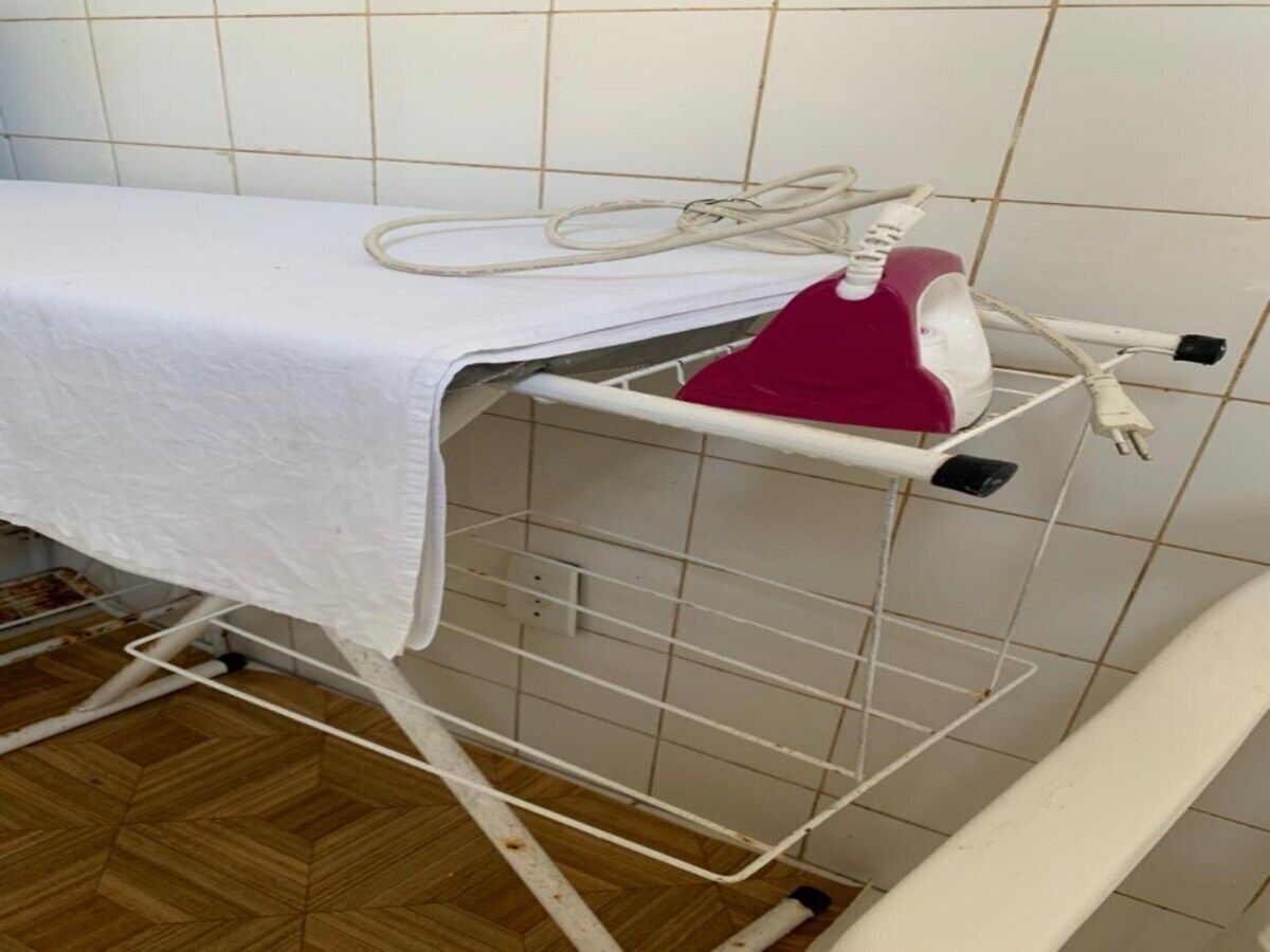 A white ironing board with a red iron and a white cloth draped over it, placed on a tiled floor.