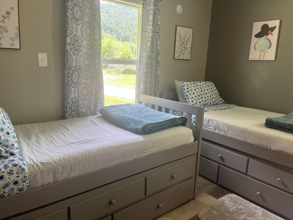 Un dormitorio con dos camas individuales, una ventana con cortinas estampadas, una lámpara de pared y un cuadro enmarcado.