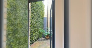 A narrow outdoor corridor with a brick wall on one side and a dense green hedge on the other. There are potted plants and a small garden area visible. - Modern 1 Bed | Private Patio | WiFi | East London (London)