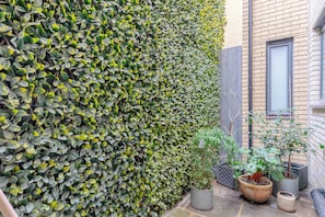 Ein Gartenbereich mit einer hohen Hecke, Kübelpflanzen und einem Backsteinhaus.