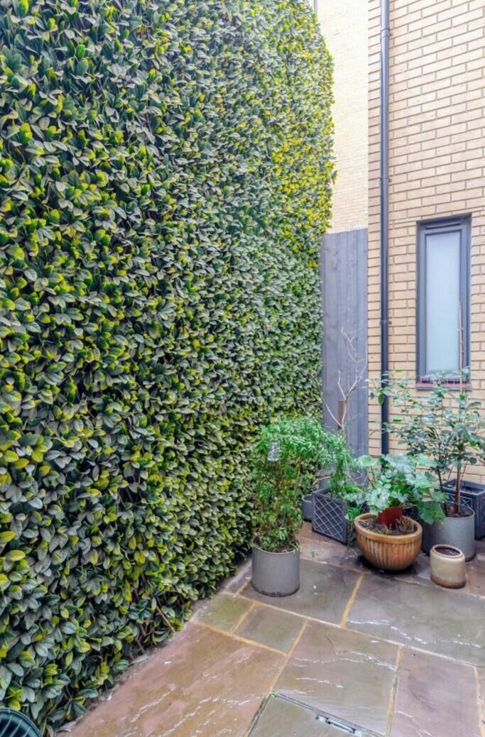 Un bâtiment en briques, avec une haie haute, des plantes en pot et une terrasse en pierre.