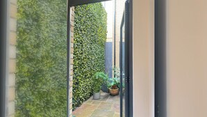 A narrow outdoor corridor with a brick wall on one side and a dense green hedge on the other. There are potted plants and a small garden area visible. - Modern 1 Bed | Private Patio | WiFi | East London (London)
