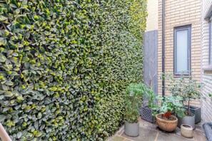 A garden area with a tall hedge, potted plants, and a brick building. - Modern 1 Bed | Private Patio | WiFi | East London (London)
