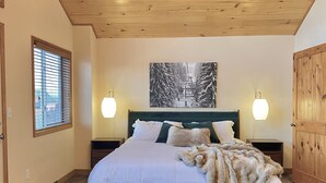 A bedroom with a bed, two bedside tables, a window with blinds, a wall-mounted painting, and a wooden door. - Pet Friendly cabin w/ fireplace and mountain views (Jefferson)