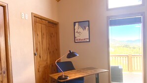 A room with a wooden door, a framed picture on the wall, a desk with a chair, and a large window with a view of a deck and mountains. - Pet Friendly cabin w/ fireplace and mountain views (Jefferson)