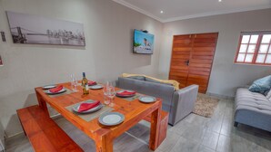 A dining area with a wooden table set for four, a grey sofa, a wooden wardrobe, and a television on the wall. - Tembo Guest Lodge  (Mbombela)