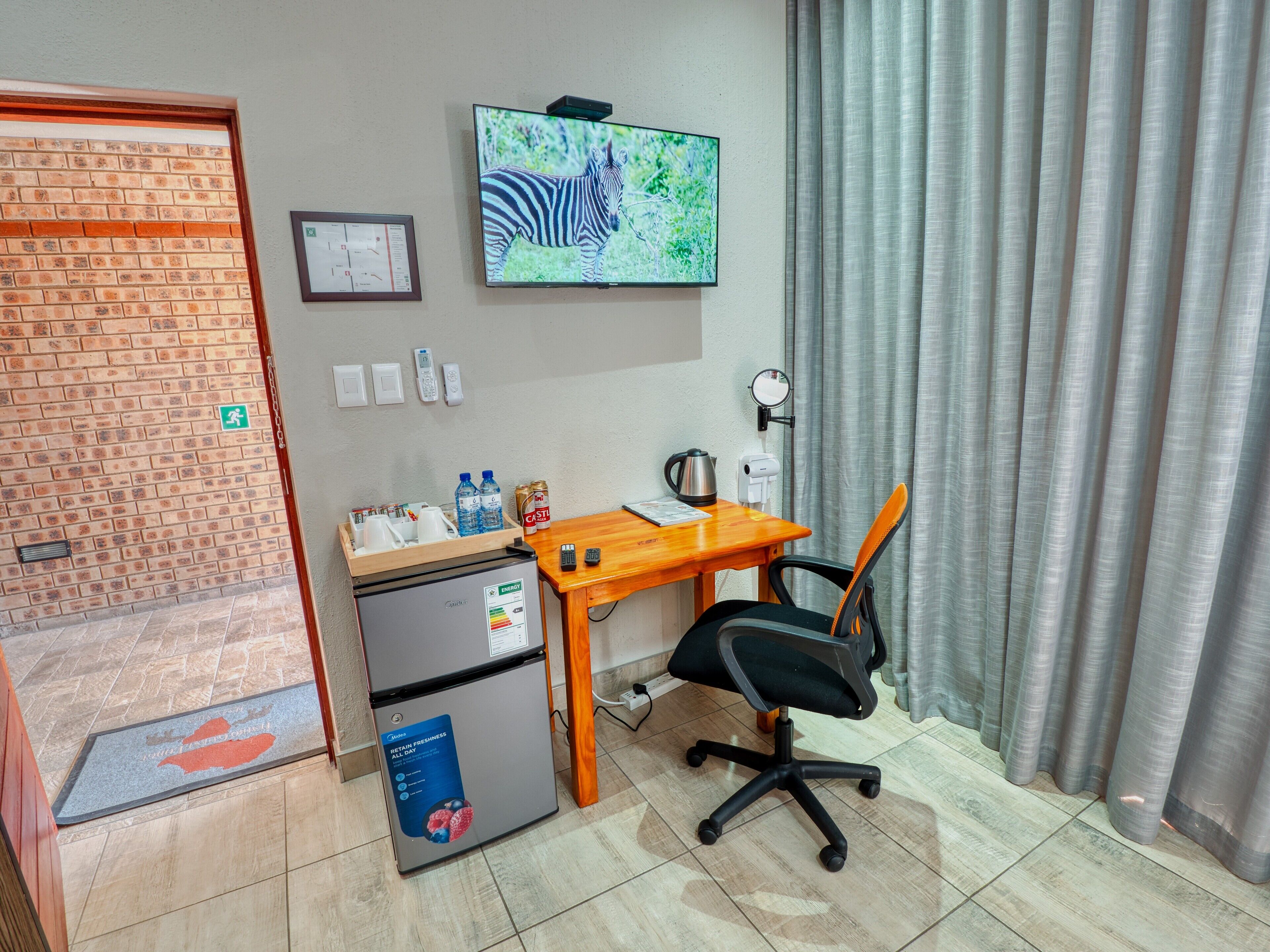 A compact hotel room with a wooden desk, a black office chair, a refrigerator, a small table with beverages and a kettle, a television mounted on the wall, and a window with curtains.
