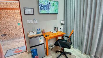 A compact hotel room with a wooden desk, a black office chair, a refrigerator, a small table with beverages and a kettle, a television mounted on the wall, and a window with curtains.