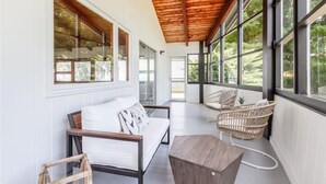 A spacious balcony with a wooden ceiling, white walls, and large windows offering a view of greenery. - Lakefront Midcentury Cottage (Williamson)