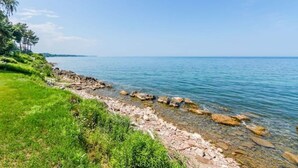 A coastal landscape with a rocky shoreline, clear blue water, and green vegetation. - Lakefront Midcentury Cottage (Williamson)