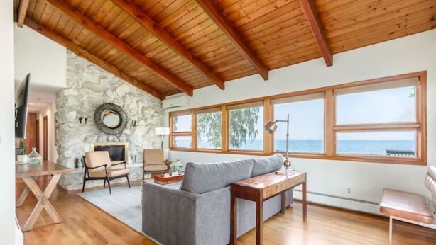 A cozy living room with a stone wall, wooden ceiling beams, a gray sofa, a wooden coffee table, and large windows overlooking the sea. - Lakefront Midcentury Cottage (Williamson)