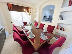 Une salle à manger avec une table en bois dressée pour six personnes, des chaises à l’assise rembourrée de tissu rouge, un lustre et une vue sur la cuisine à travers une arche.