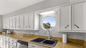 A kitchen with white cabinets, a granite countertop, a double sink, and a window with a view of palm trees. - MultiDeck VIEWS *Sleeps 14* Pool & PingPong Table *Games Galore* *Fire-Pit* (Galveston)