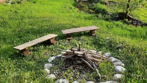 A serene outdoor setting with a circular stone fire pit surrounded by rocks and logs, two wooden benches, and lush greenery. - In the middle of nature, an oasis of peace
 (Log Cezsoški)