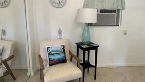 Een kamer met een stoel, een klein tafeltje met een lamp, een klok en een raam met gordijn.