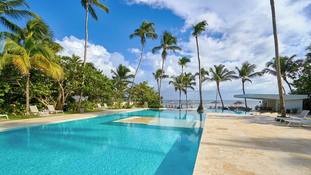Una piscina azul cristalina rodeada de palmeras y sillones de descanso, con vista al océano.