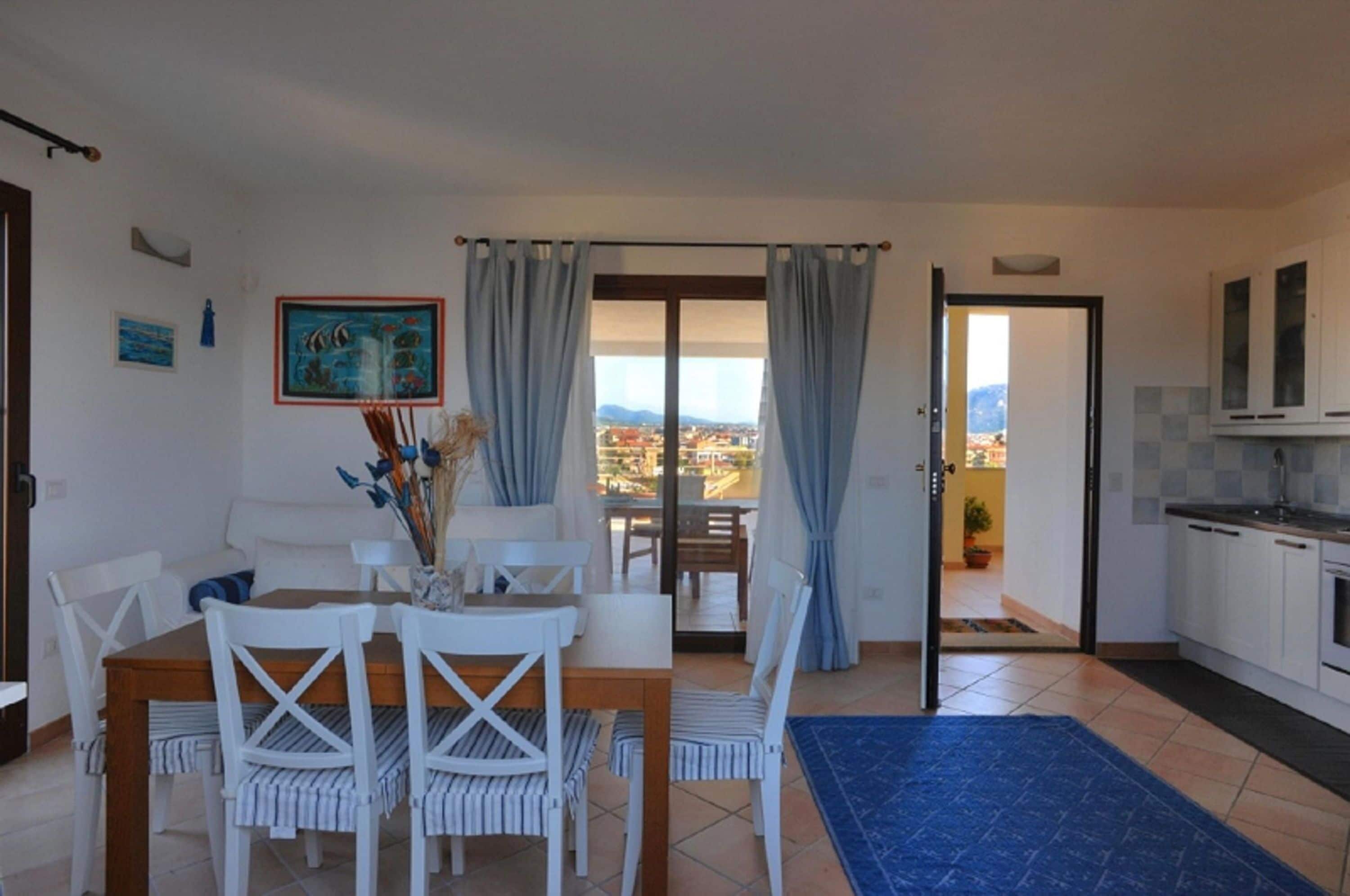A dining area with a table and chairs, a view through a glass door, and a kitchen with cabinets and a sink.